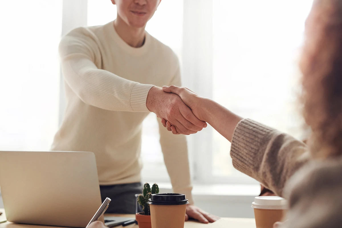 One person stands and shakes hands with another person sitting across a table. A laptop and two coffee cups are pictured on the table.
