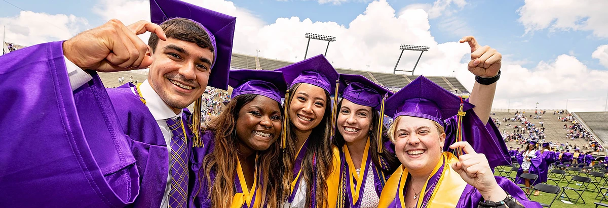 5 smiling individuals in purple caps and gowns in a stadium