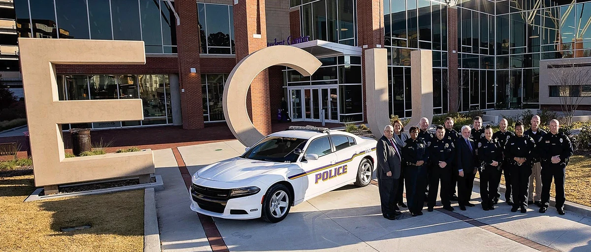 A group of police officers and officials stand beside a police car in front of a building with large "ECU" letters.