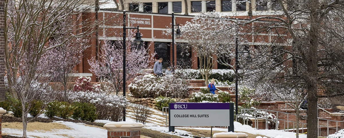 A snowy scene outside College Hill Suites at ECU, with flowering trees and people walking nearby.