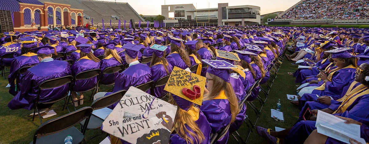 A large group of graduates in purple gowns and decorated caps are seated at an outdoor commencement ceremony in a stadium.