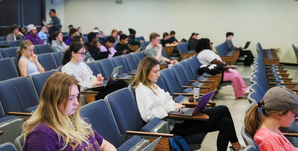 A group of students is sitting in a lecture hall, many using laptops.