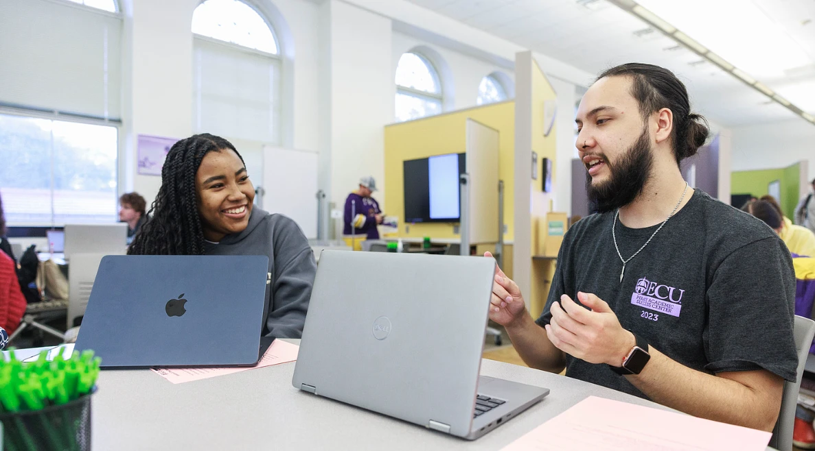 Two people are smiling and working on laptops in a classroom setting.