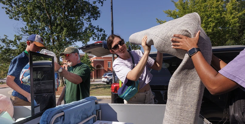 A group of people are unloading items from a car, including a rug, in a sunny outdoor setting.