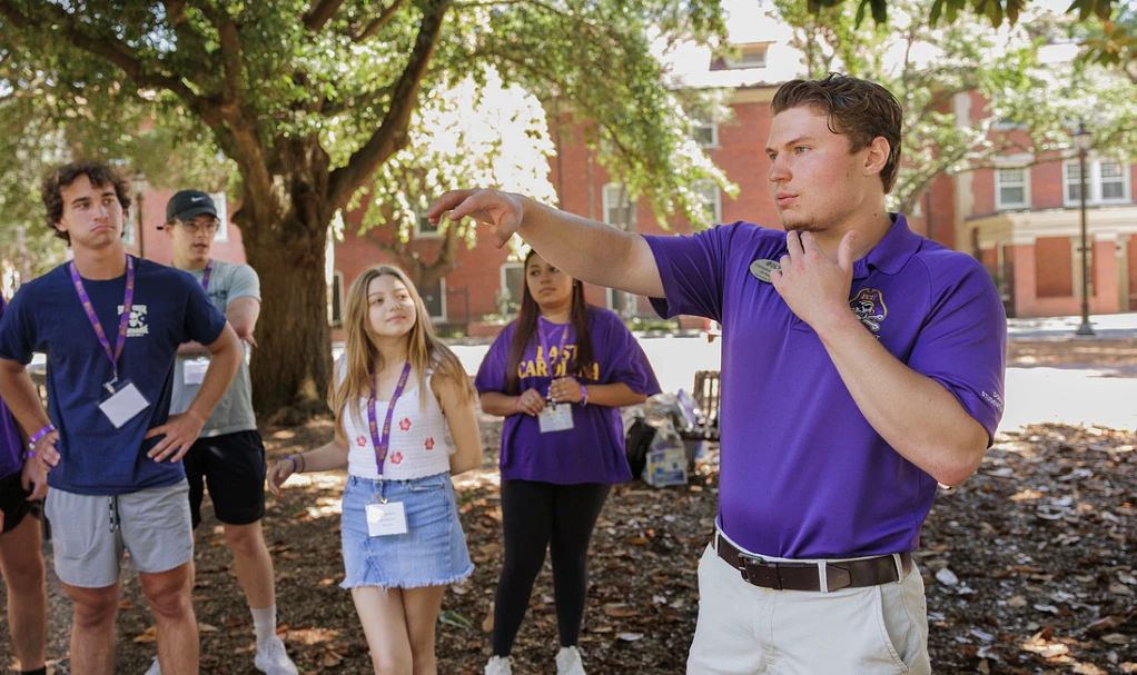 A young man in a purple shirt is guiding a group of people outdoors, possibly during a tour or orientation.