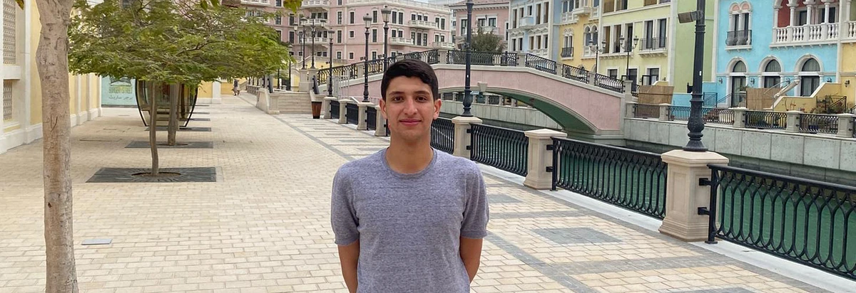 Student in grey shirt and arms behind his back on a sidewalk with a pink bridge in the background